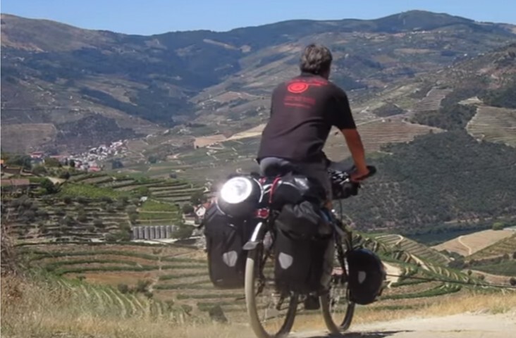 De fietsende kok Peter trekt met zijn volgepakte fiets over een zandpad door een uitgestrekt, terrasvormig berglandschap met wijngaarden en zonlicht.